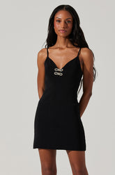 A woman is wearing a black sleeveless dress with thin spaghetti straps. The dress features a V-neckline with decorative silver embellishments at the center of the bust. The dress has a fitted silhouette and falls to mid-thigh length.